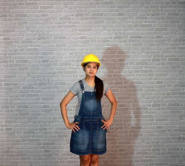 Teknisyen kadın gri tişörtlü ve kot önlüklü beyaz kasklı gri tuğla desenli arka planda akimbo ayakta duran kadın..