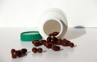 Beyaz zeminde koyu kahverengi ilaç yığını ve yeşil kapaklı beyaz plastik şişe..