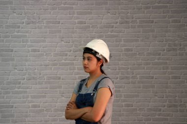 Teknisyen kadın, gri tişörtlü ve kot önlüklü beyaz kasklı 45 derecelik açıda duran ve gri tuğla desenli arka planda göğsüne sarılan bir kadın giysisi giyiyor..