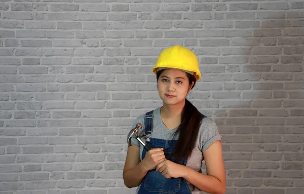 女技师用黄色头盔装饰灰色T恤衫 牛仔围裙站立 双手拿铁锤在灰色砖图案背景上 — 图库照片