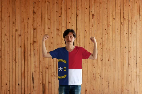 ノースカロライナ州の旗色のシャツを着て 木製の壁の背景に両方の拳を上げて立っている男 アメリカの州 — ストック写真
