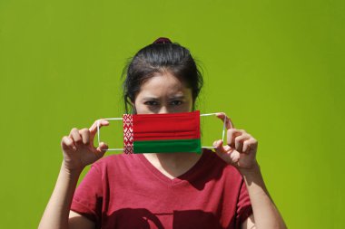 Elinde Beyaz Rusya bayrağı deseni olan bir kadın ve hijyenik maske yeşil arka planda yüzünü kapatmak için kaldırıyor. Maske, Minik Parçacık ya da virüs koronasına karşı çok iyi bir korumadır..