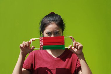 A woman and hygienic mask with Belarus flag pattern in her hand and raises it to cover her face on green background. A mask is a very good protection from Tiny Particle or virus corona. clipart