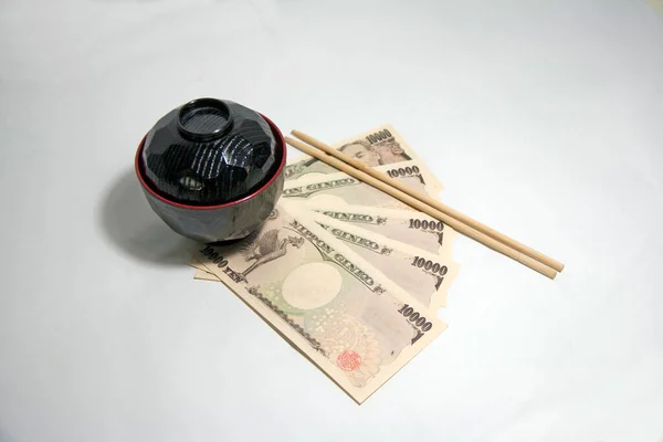Black color red edge Miso soup bowl and wooden Chopsticks with Yen banknotes of Japan on the white background. The concept of Eat well live happily by Japanese style.