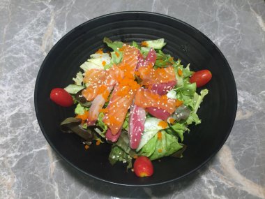 Mermer masadaki siyah kasede saşimi salatası, çiğ balık salatası ve domates, üzerine beyaz susam serpiştirilmiş..