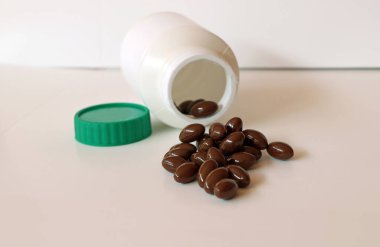 Beyaz zeminde koyu kahverengi ilaç yığını ve yeşil kapaklı beyaz plastik şişe..