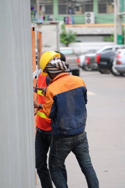 Pathumthani, Tayland - 6 Ağustos 2017: Turuncu ve mavi elbiseli iki inşaat işçisi yolun kenarındaki çinko duvarı elektrikli matkapla deliyorlar..