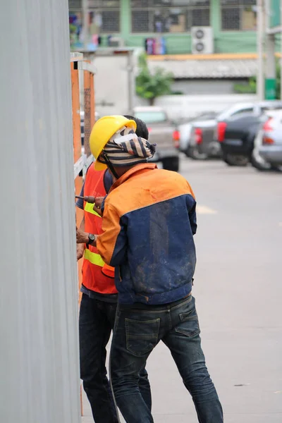 泰国帕通塔尼 2017年8月6日 两名身穿橙色和蓝色衣服的建筑工人在路边用电钻钻钻钻锌墙 — 图库照片