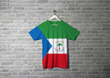 Tişörtünde ve duvarında tuğla desenli duvar kâğıdı asılı Ekvator Gine bayrağı, mavi üçgenli yeşil beyaz ve kırmızı ve Ulusal armalı..