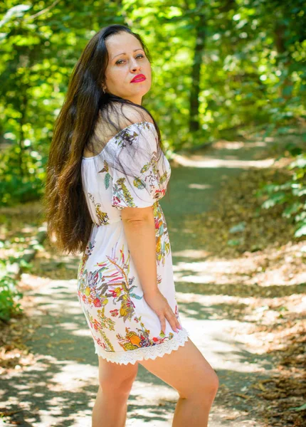 A middle-aged woman of European appearance with long dark hair in a summer dress is resting in the city\'s park. A woman a little overweight without complexes