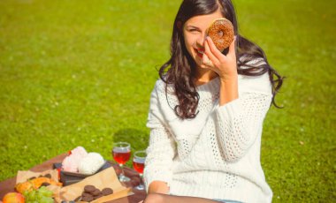 Beyaz örgü kazaklı, güzel esmer bir kadın piknikte lezzetli bir çikolatalı çörek yiyor ve bugünün tadını çıkarıyor. Fast food 'un artıları ve eksileri. kadın ve diyet