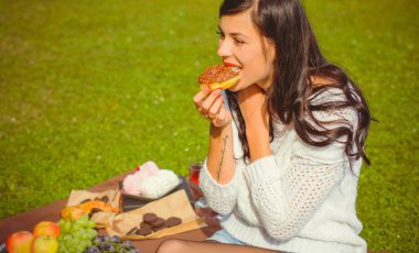 Beyaz örgü kazaklı, güzel esmer bir kadın piknikte lezzetli bir çikolatalı çörek yiyor ve bugünün tadını çıkarıyor. Fast food 'un artıları ve eksileri. kadın ve diyet