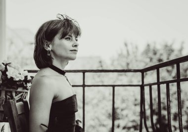 Siyah, eski moda uzun elbiseli, şık aksesuarlı, kolyeli, küpeli ve taşlı yüzüklü orta yaşlı güzel bir kadın. Siyah elbiseli kadın ve balkondaki boa