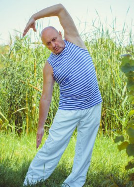 Kıdemli adam doğa üzerinde egzersiz yapar. Fizyoterapi konsepti. Sokakta spor yapan yaşlı bir adam. Sazlıklara karşı egzersiz yaparken kaslarını esneten olgun bir adam..