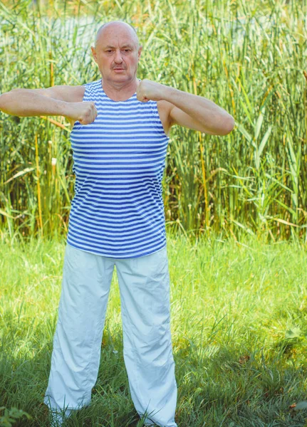 Senior man do exercises on a nature. Physiotherapy concept. Elderly man practicing sports on the street. Full length of mature man stretching muscles during exercise against reeds.