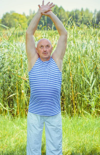 Senior man do exercises on a nature. Physiotherapy concept. Elderly man practicing sports on the street. Full length of mature man stretching muscles during exercise against reeds.