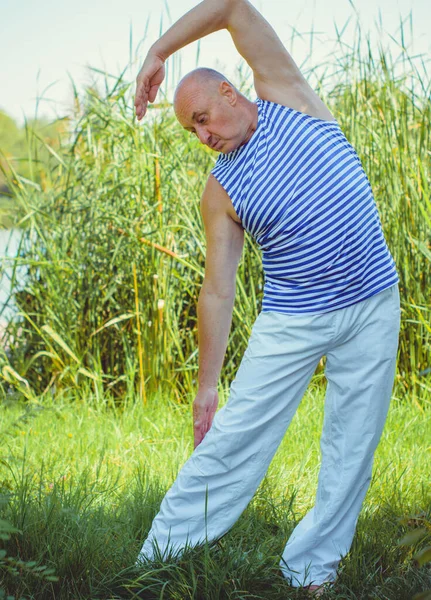 Senior man do exercises on a nature. Physiotherapy concept. Elderly man practicing sports on the street. Full length of mature man stretching muscles during exercise against reeds.