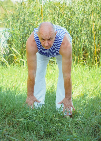 Senior man do exercises on a nature. Physiotherapy concept. Elderly man practicing sports on the street. Full length of mature man stretching muscles during exercise against reeds.