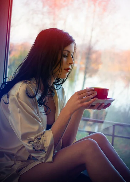 若い美しい夢のブルネットの女性は窓のそばに家に座ってランジェリーを着てコーヒーを飲む 庭の木の背景 女性の夢と願いの概念 — ストック写真