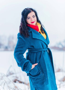 Kış tatili konsepti. Tarz sahibi Arap kadın dışarıda karlı bir güne bakıyor. Mavi alpaka paltolu kadın ve kış günü açık havada kırmızı-sarı atkı.