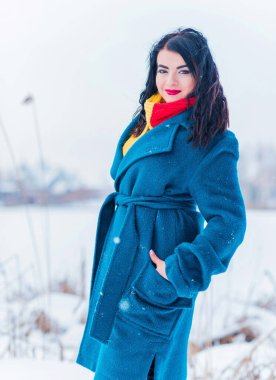 Kış tatili konsepti. Tarz sahibi Arap kadın dışarıda karlı bir güne bakıyor. Mavi alpaka paltolu kadın ve kış günü açık havada kırmızı-sarı atkı.