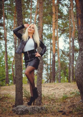 Ormanda genç ve seksi bir kadın deri ceket giyiyor ve etek kendini gösteriyor ve ormanda çam ağacının arka planına karşı elbiseler giyiyor.