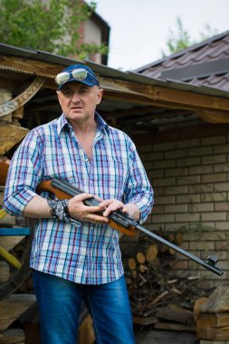 Adam, odunların yanında silahı ve yakacak odunu olan ekose gömlekli, beysbol şapkalı ve erkek aksesuarlarıyla şık görünümlü gözlüklü bir Amerikalı.