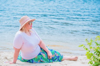 Artı plajda bir model. Şişman kadın hayattan zevk alıyor. Nehir kenarındaki doğada, kumların üzerinde dinlenen güzel bir kadın. Kadın hayatı kavramı.