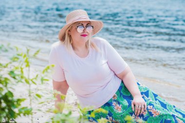 Artı plajda bir model. Şişman kadın hayattan zevk alıyor. Nehir kenarındaki doğada, kumların üzerinde dinlenen güzel bir kadın. Kadın hayatı kavramı.