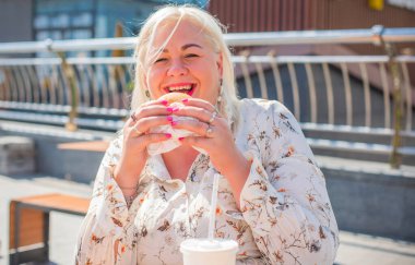 Tombul, güzel, mutlu, iri yarı bir kadın açık havada iyi bir ruh hali içinde hamburger yer, diyetten vazgeçer, mutlu bir hayat sürer ve vücuda pozitif bir konsept yaratır.