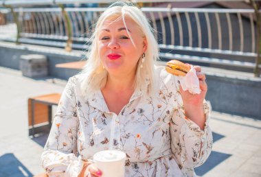 Tombul, güzel, mutlu, iri yarı bir kadın açık havada iyi bir ruh hali içinde hamburger yer, diyetten vazgeçer, mutlu bir hayat sürer ve vücuda pozitif bir konsept yaratır.