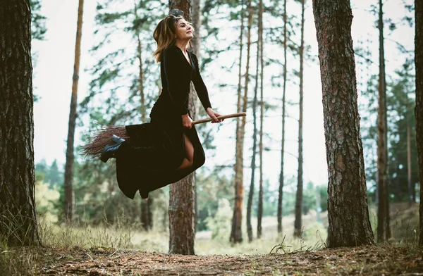 若い美しく神秘的な魔女の女性が森の中のブルームを飛んでいます ほうきの上の魔女 無料のスペース ハロウィーンの休日魔法 — ストック写真
