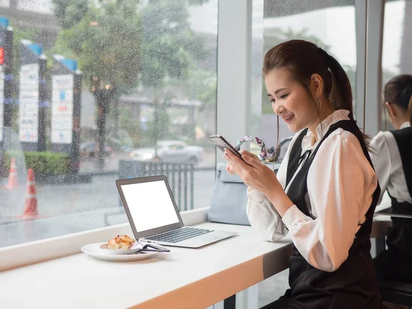 Side view of Asia young business woman in white shirt using smartphone and laptop sitting near window outside raining and uses laptop and smartphone
