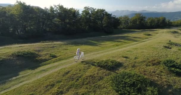 Tiro de uma jovem mulherem um vestido leve e chapéu andar de bicicleta na estrada rural — Vídeo de Stock