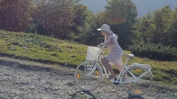 Tiro de uma jovem mulherem um vestido leve e chapéu andar de bicicleta na estrada rural — Vídeo de Stock