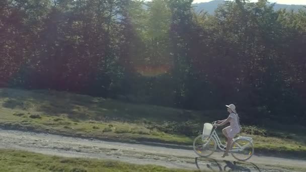 Снимок молодой женщины в светлом платье и велосипеде на шляпе по сельской дороге — стоковое видео