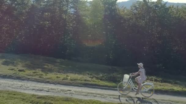 Foto van een jonge vrouw in een lichte jurk en hoed paardrijden fiets op het platteland weg — Stockvideo