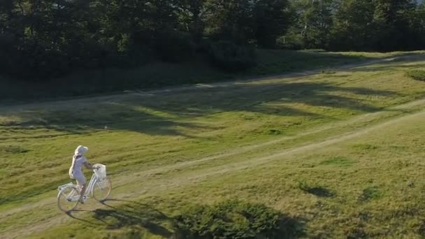 Aufnahme einer jungen Frau in hellem Kleid und Hut, die auf einer Landstraße Fahrrad fährt — Stockvideo