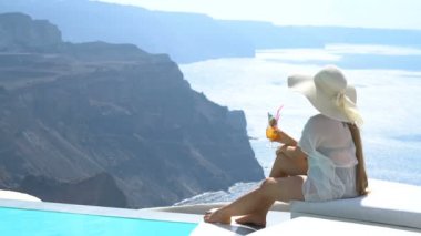 Genç kadın havuz kenarında Santorini büyüleyici bir manzarasına sahip bir kokteyl içme