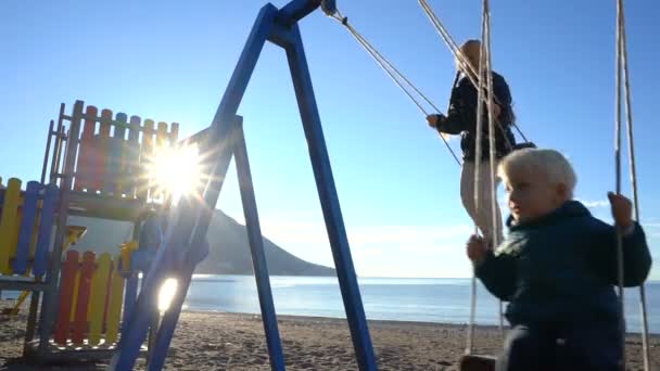 Un niño de tres años y una niña de seis años balanceándose en un columpio en una mañana soleada en la costa — Vídeo de stock