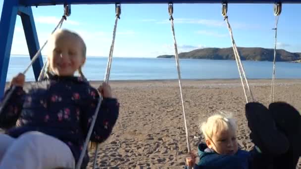 在海岸阳光明媚的早晨, 一名3岁的男孩和一名6岁的女孩在秋千上荡秋千 — 图库视频影像