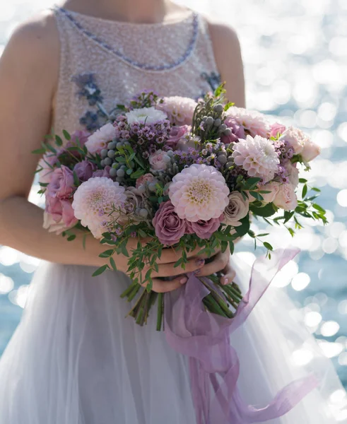 La novia sostiene un ramo de boda rosa y lila en sus brazos contra el fondo del mar — Foto de Stock