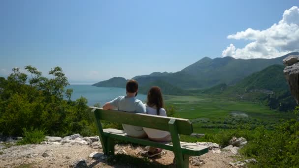Пара сидит на скамейке и любуется видом на озеро и холмы — стоковое видео