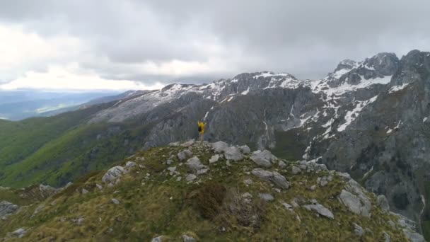 Um caminhante fica no topo de uma montanha e olha para montanhas cobertas de neve, levantando as mãos em gesto de vitória . — Vídeo de Stock