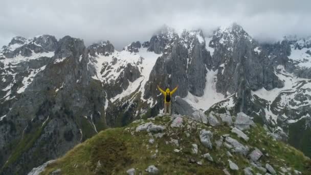 Hiker stoi na szczycie góry i patrzy na ośnieżone góry, podnosząc ręce w gestu zwycięstwa. — Wideo stockowe