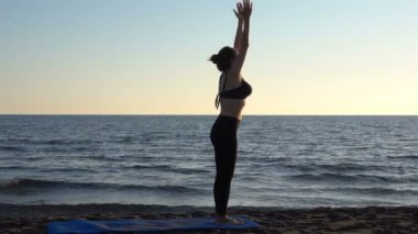 Sakin denizin yanındaki sahilde yoga yapan beyaz bir kadın, gündoğumu geçmişi var..