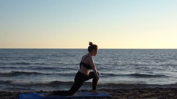 Młoda kaukaska kobieta ćwiczy jogę na plaży w pobliżu spokojnego morza, tło wschodu słońca. — Wideo stockowe