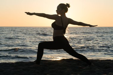 Genç beyaz kadın sakin denizin yanındaki sahilde yoga yapıyor.