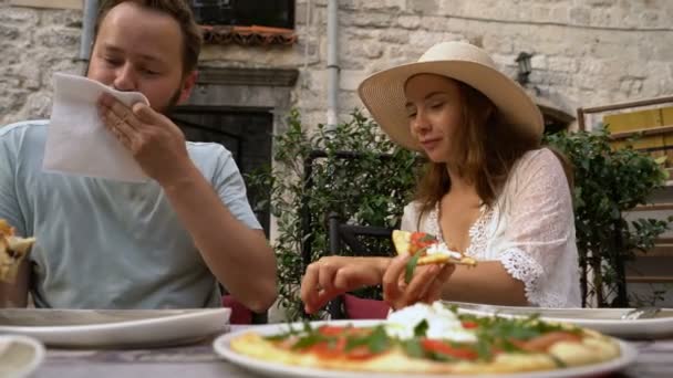 Молодая пара сидит в кафе, ест пиццу, разговаривает и улыбается — стоковое видео
