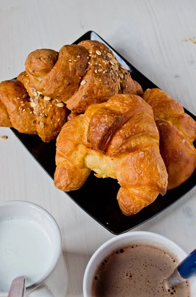 Tereyağı ve mısır gevreği ile mükemmel sağlıklı kahvaltı siyah tepside kruvasan ve ağarmış ahşap masa ile bir fincan süt ve çikolata.